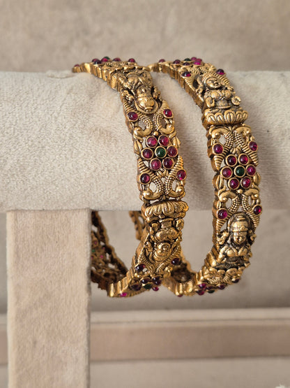 Two ornate gold-toned bangles with detailed relief carvings and small red and green stone accents, by naitri.in