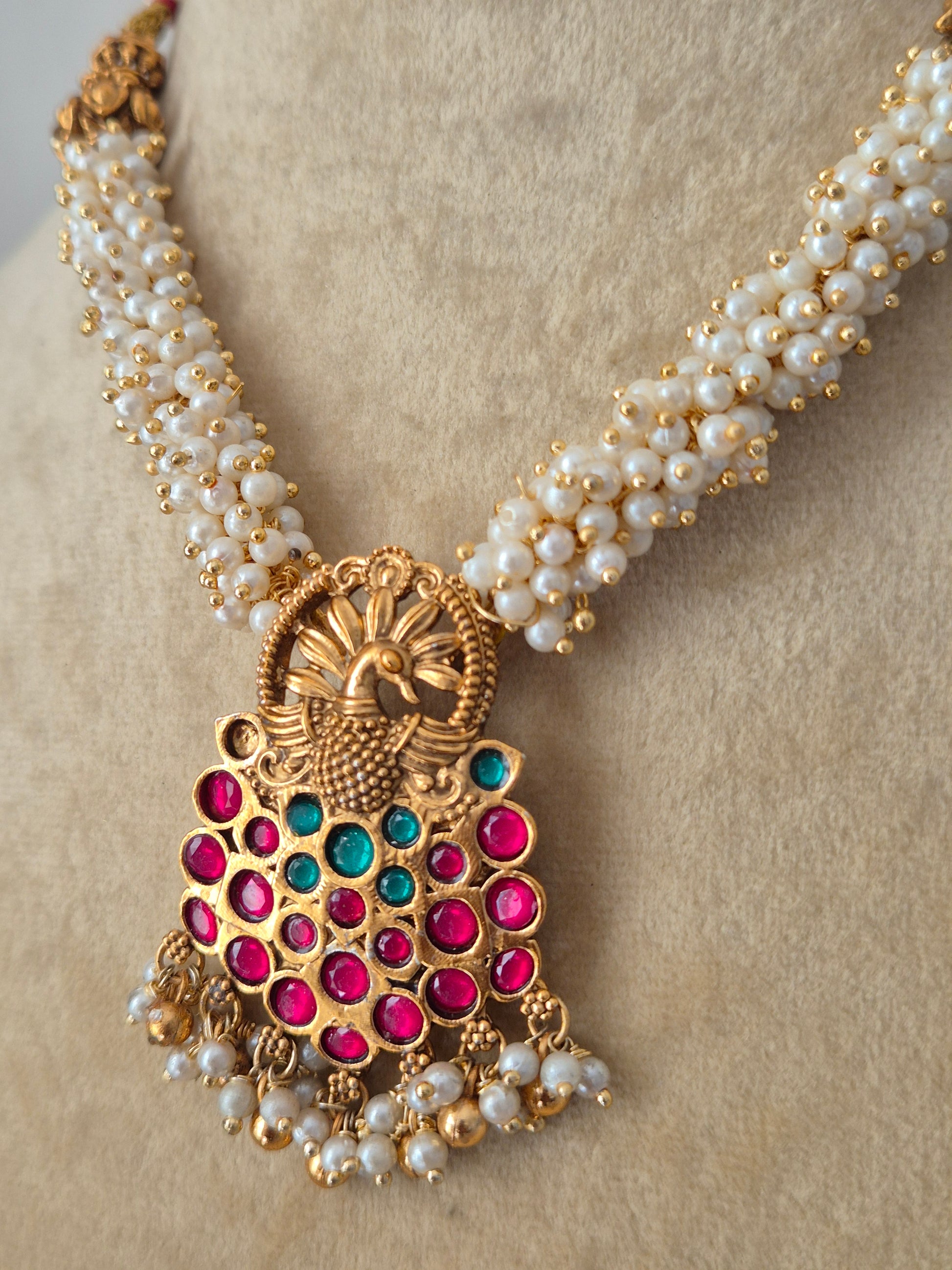 Traditional Indian gold-polished brass necklace set showcasing an intricate antique filigree pendant with ruby-red and emerald-green gemstones, suspended from a woven pearl strand by naitri.in