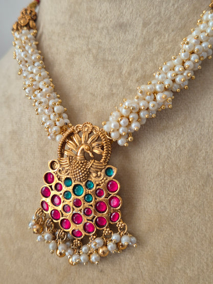 Traditional Indian gold-polished brass necklace set showcasing an intricate antique filigree pendant with ruby-red and emerald-green gemstones, suspended from a woven pearl strand by naitri.in