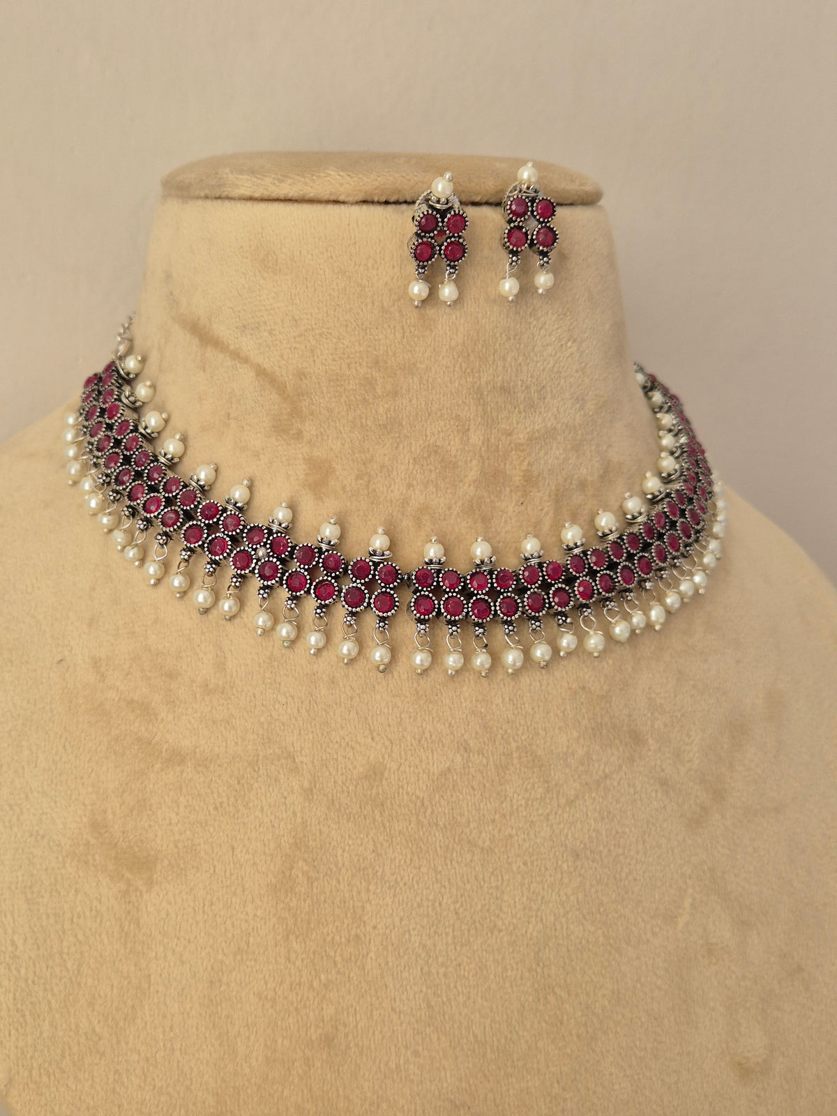 A traditional Indian jewelry set featuring a choker necklace and matching earrings. The jewelry features silver-toned metalwork, deep pinkish-red stones, and white pearl bead accents by naitri.in