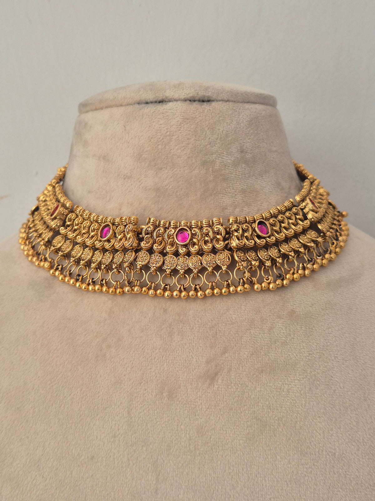 Traditional gold choker necklace featuring intricate antique detailing with pink stone accents and small dangling beads, with matching bell-shaped jhumka earrings by naitri.in