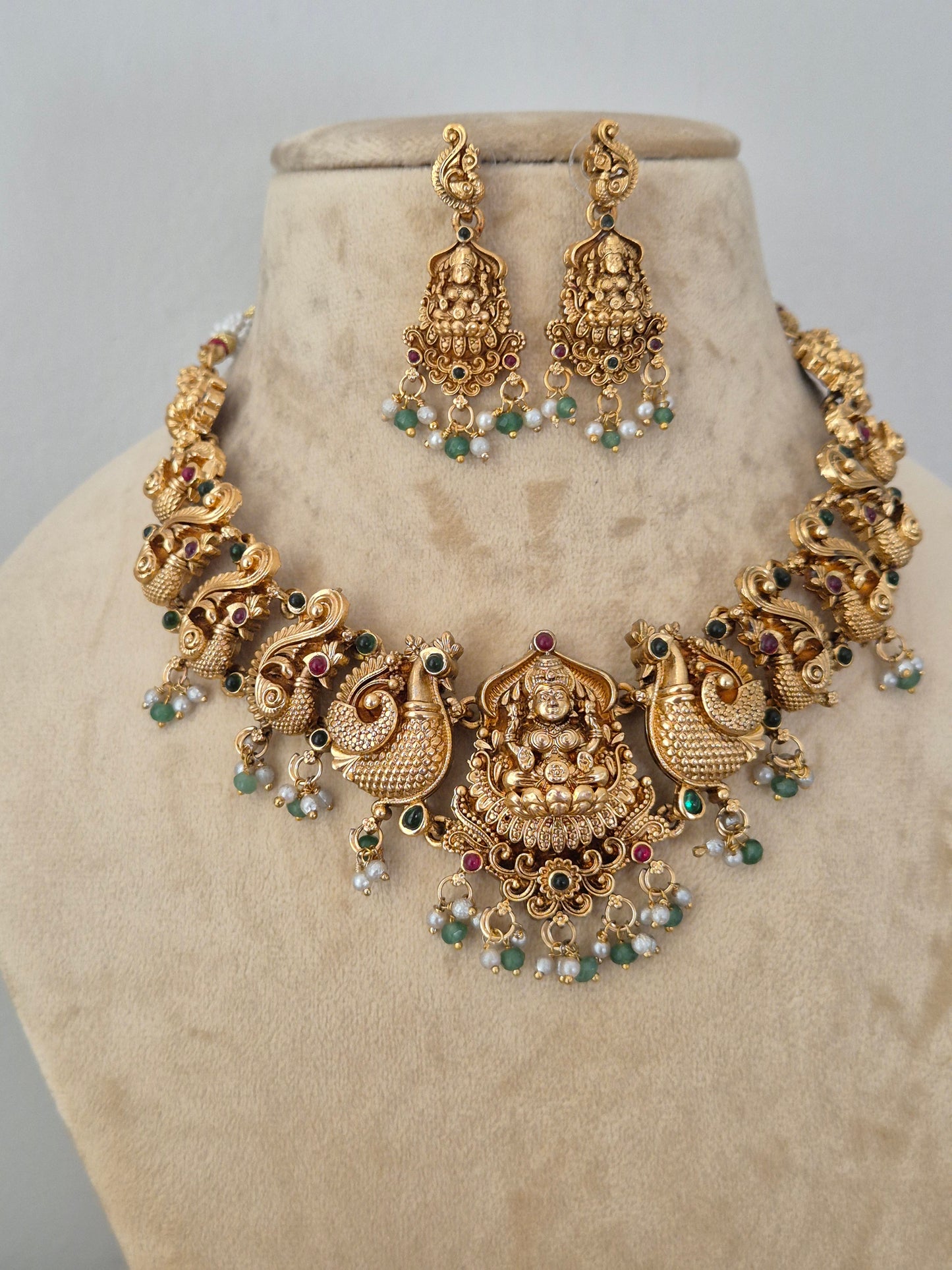 Gold-toned traditional Indian jewelry set featuring a choker necklace and matching earrings, decorated with detailed motifs, small green and red stones, and white pearl-like beads, by naitri.in