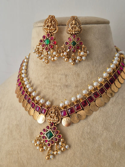 Traditional Indian temple jewelry set in polished brass, showcasing an antique finish pendant with red and green gemstone accents, a layered pearl band, and a classic coin fringe by naitri.in