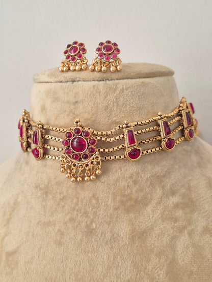 Antique gold-toned choker necklace showcasing an intricate metallic floral band, vibrant red stone clusters, and delicate hanging gold bead drops, shown with coordinating round stud earrings by naitri.in