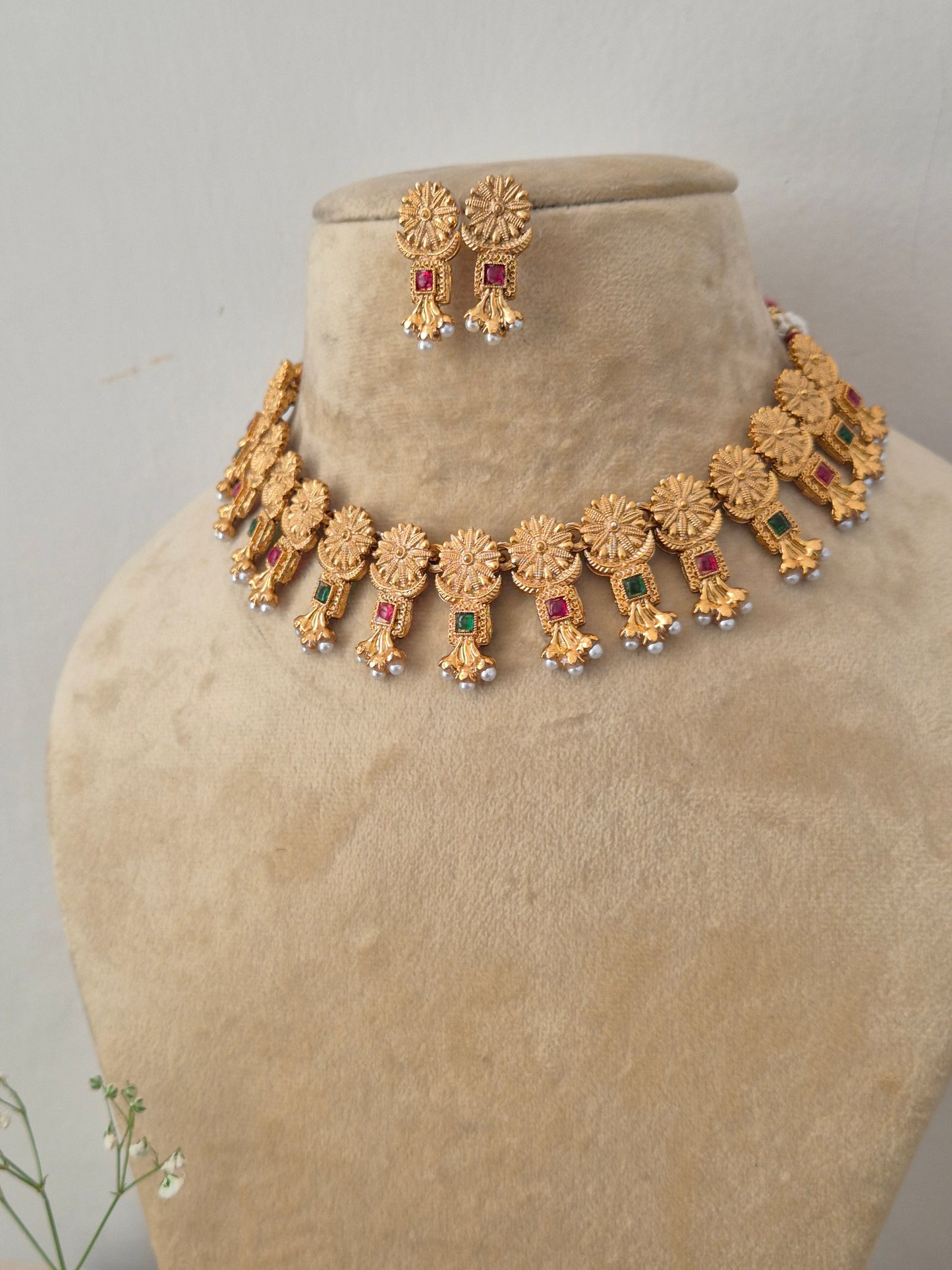 Gold-plated ethnic choker set featuring textured metallic medallions, alternating ruby-red and emerald-green stone accents, and tiny hanging pearl drops by naitri.in