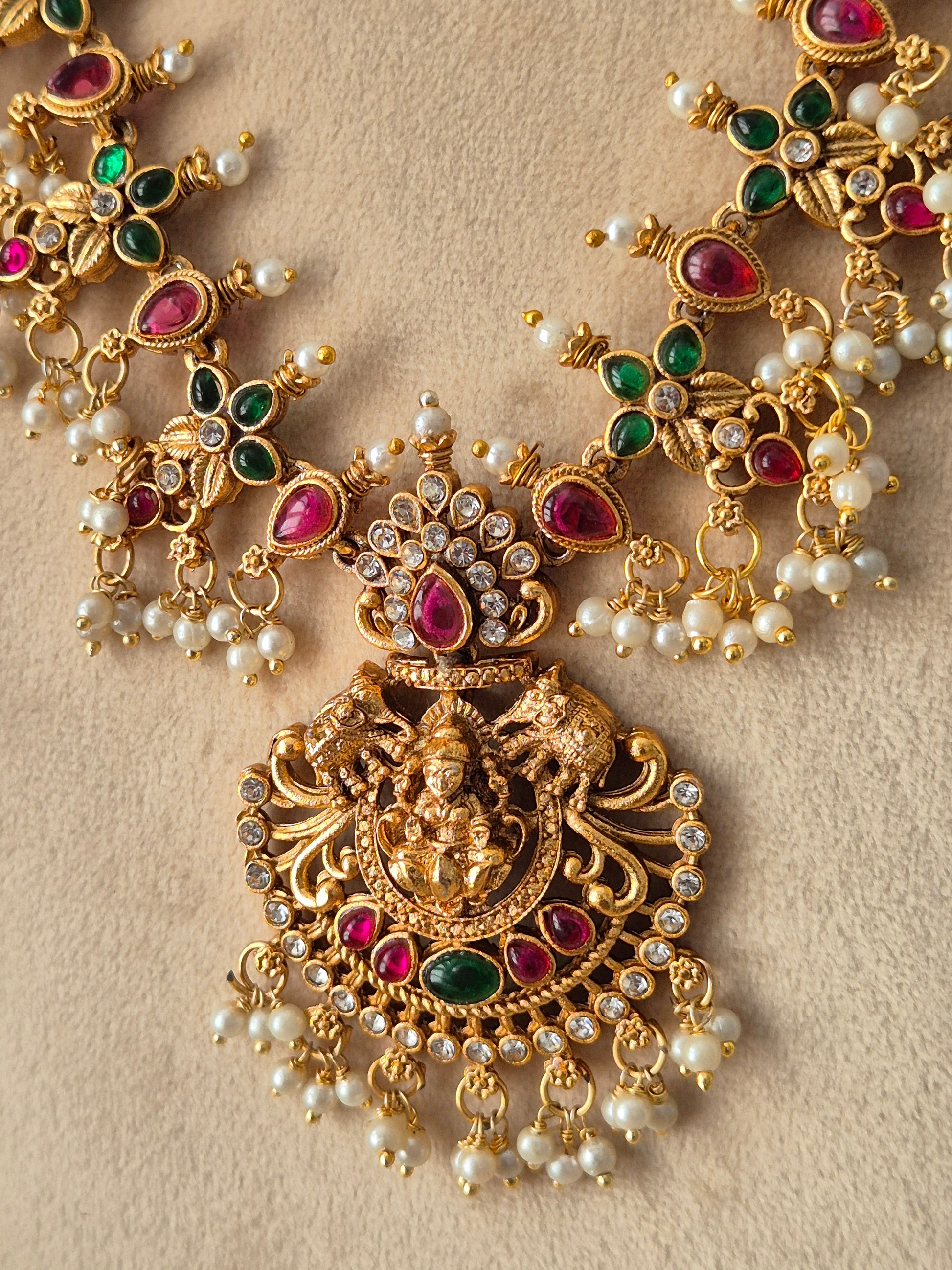 An ornate gold-toned traditional jewelry piece featuring a central deity motif, embellished with red and green teardrop-shaped stones, round clear stones, and small white pearl-like beads, by naitri.in