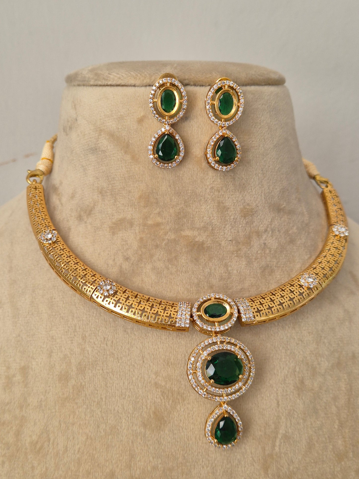 Gold-plated ethnic choker set featuring an openwork filigree band, a prominent oval-cut green stone pendant surrounded by crystals, and matching teardrop earrings by naitri.in