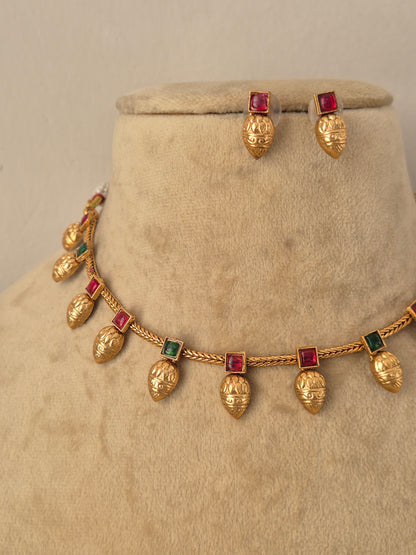 Gold jewelry set featuring a necklace and matching earrings with textured gold leaf pomegranate-shaped charms and square red-green  gemstone accents, by naitri.in