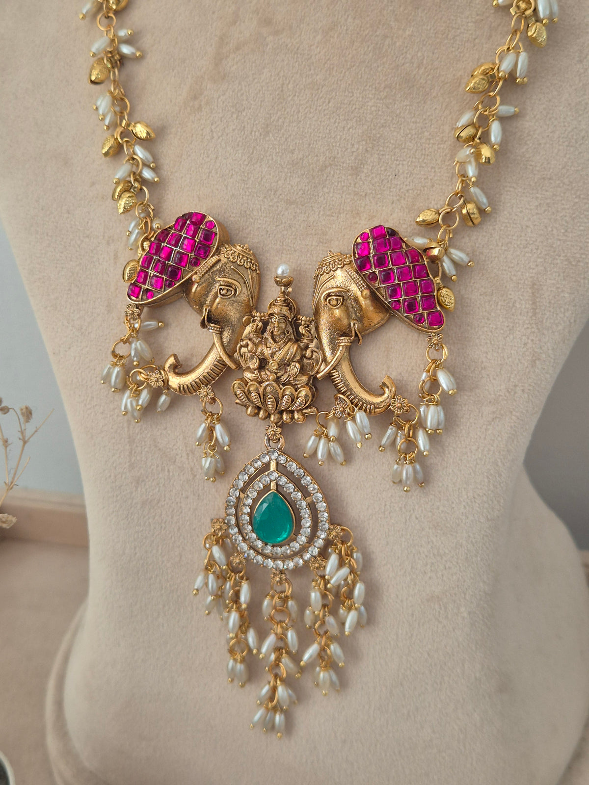 gold temple necklace featuring magenta stone-encrusted elephant motifs, a green teardrop center pendant with dangling pearls, Gold temple necklace set with pink elephant accents, a green stone pendant, and pearl drop details by naitri.in