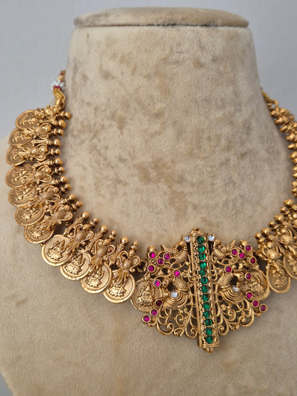 An ornate gold-toned statement necklace features intricate filigree detailing, multiple dangling coin-like elements, and a central vertical bar set with green stones and small pink stones by naitri.in