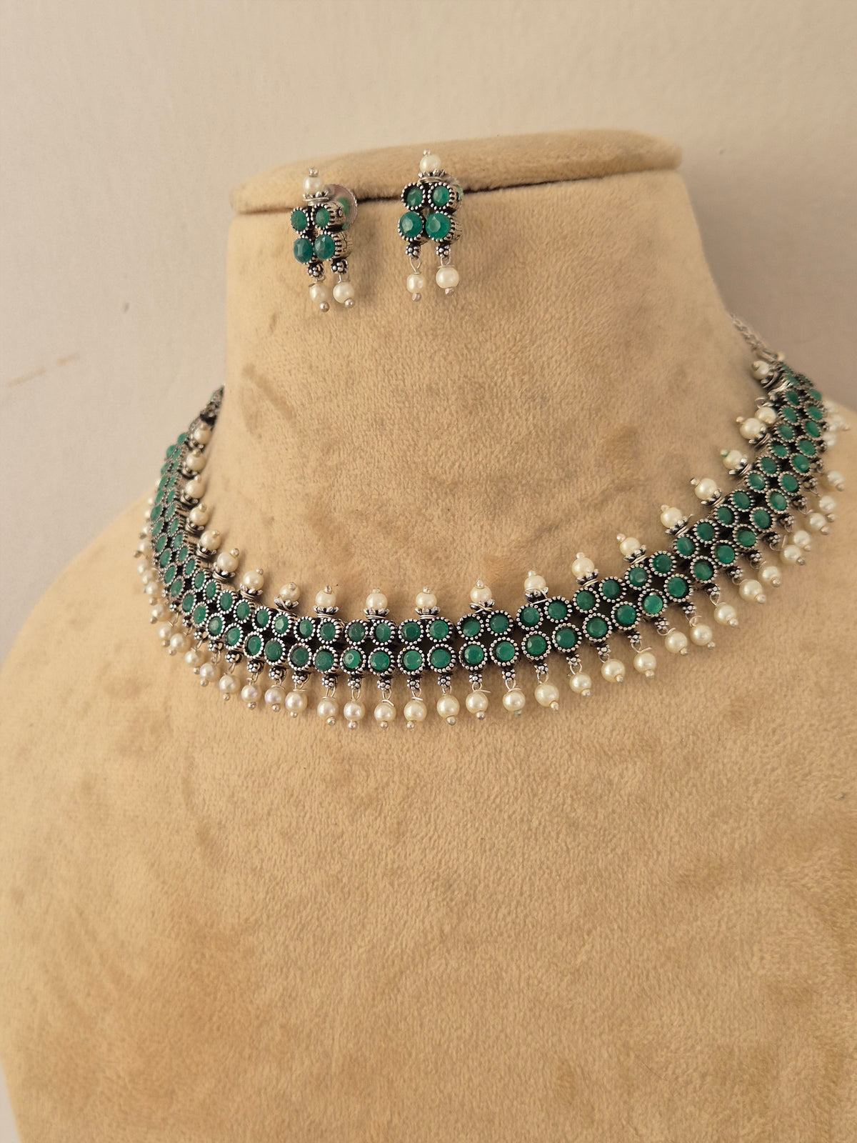 A traditional Indian jewelry set featuring a choker necklace and matching earrings, The set features silver-plated metal work, deep green stones, and white faux pearls by naitri.in