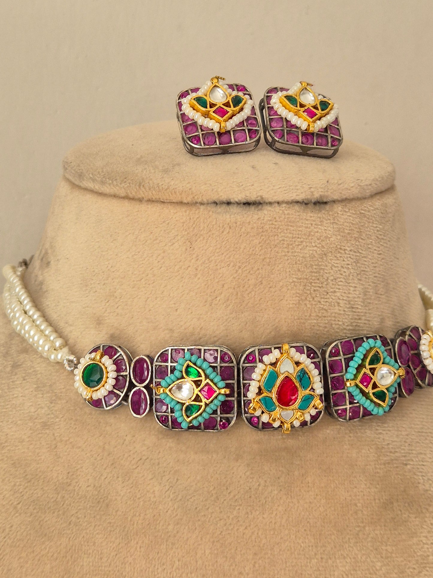 A jewelry set  featuring a statement necklace and matching earrings. The jewelry has silver-toned metal settings with purple gemstone borders, central red and turquoise stones, and white pearl-like accents by naitri.in