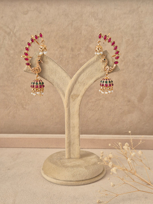 Gold-plated peacock jhumka earrings featuring a curved hoop studded with ruby-red stones leading to a bell-shaped dome with pearl drops by naitri.in