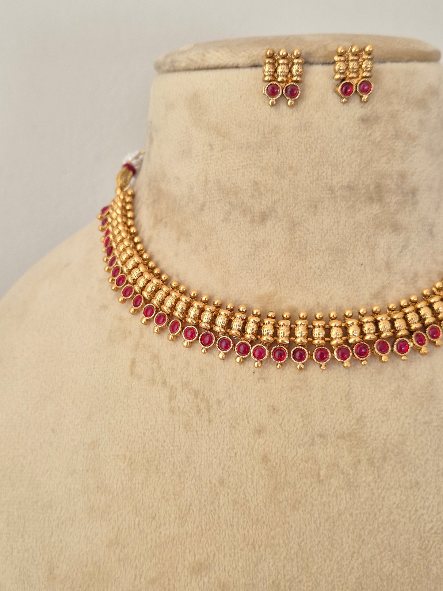 Gold-toned traditional necklace and matching earrings with red gemstone accents, necklace is designed with a row of small vertical bars leading down to a dense row of deep pink or ruby red gemstones y naitri.in