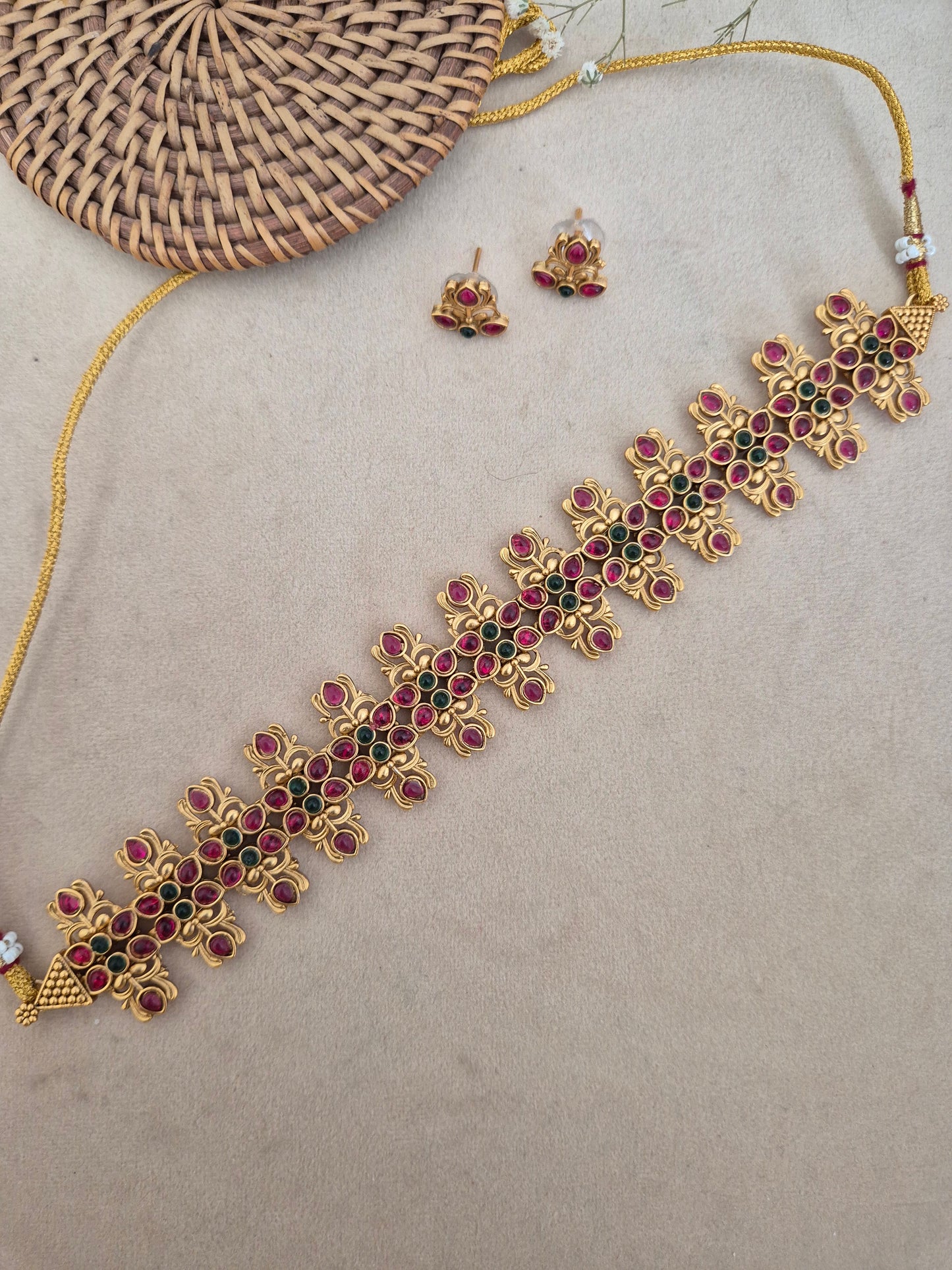 Traditional Indian antique-finish brass choker set showcasing structured rows of pinkish-red gemstones, leaf-shaped gold metalwork accents, and coordinating floral stud earrings from the Naitri collection by naitri.in