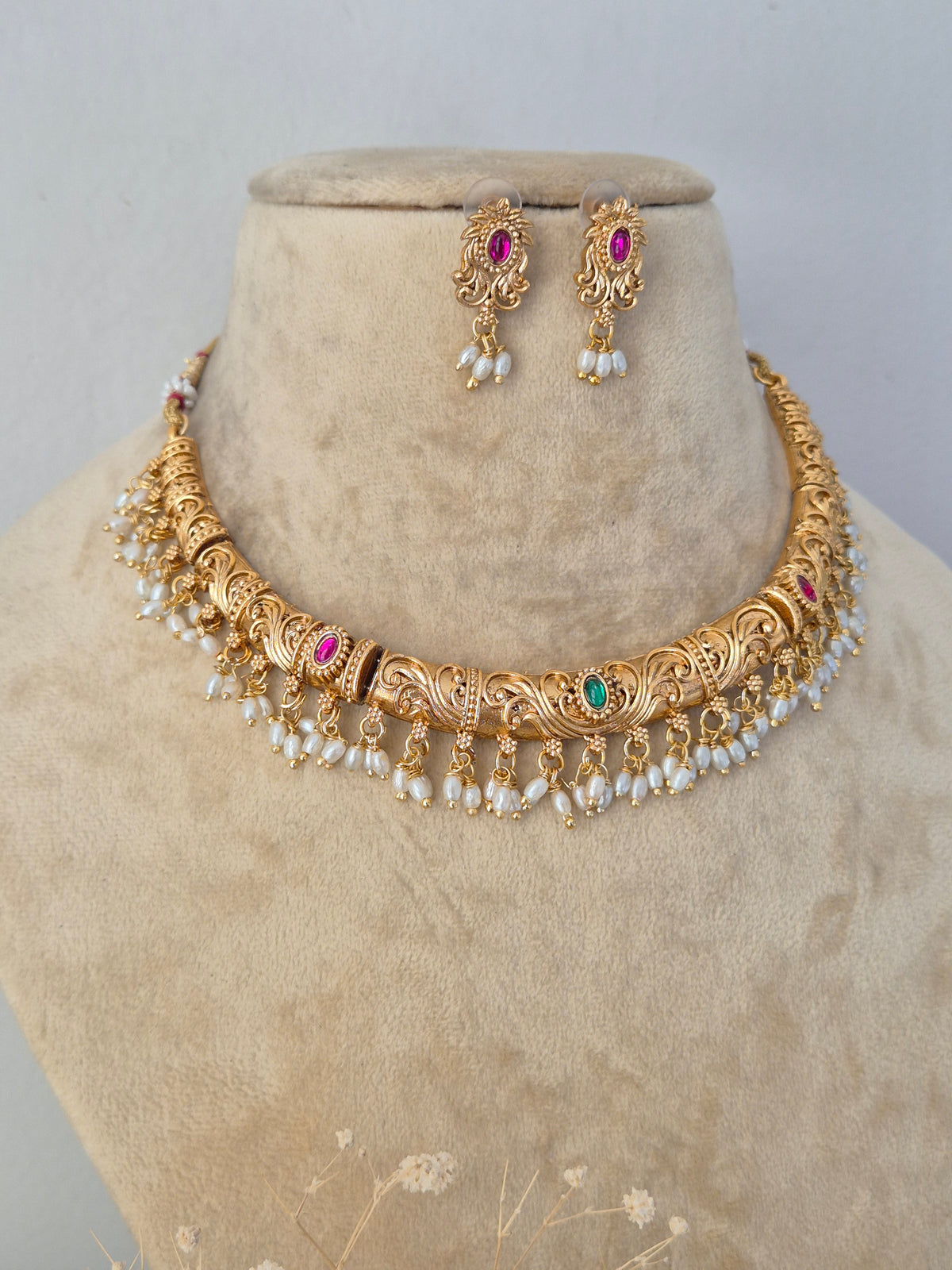 Ornate gold-toned brass alloy necklace featuring intricate traditional peacock and floral motifs embedded with red and green stones, edged with dangling white pearls, paired with matching jhumka earrings by naitri.in