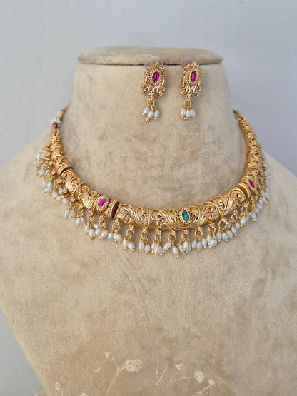 Ornate gold-toned brass alloy necklace featuring intricate traditional peacock and floral motifs embedded with red and green stones, edged with dangling white pearls, paired with matching jhumka earrings by naitri.in
