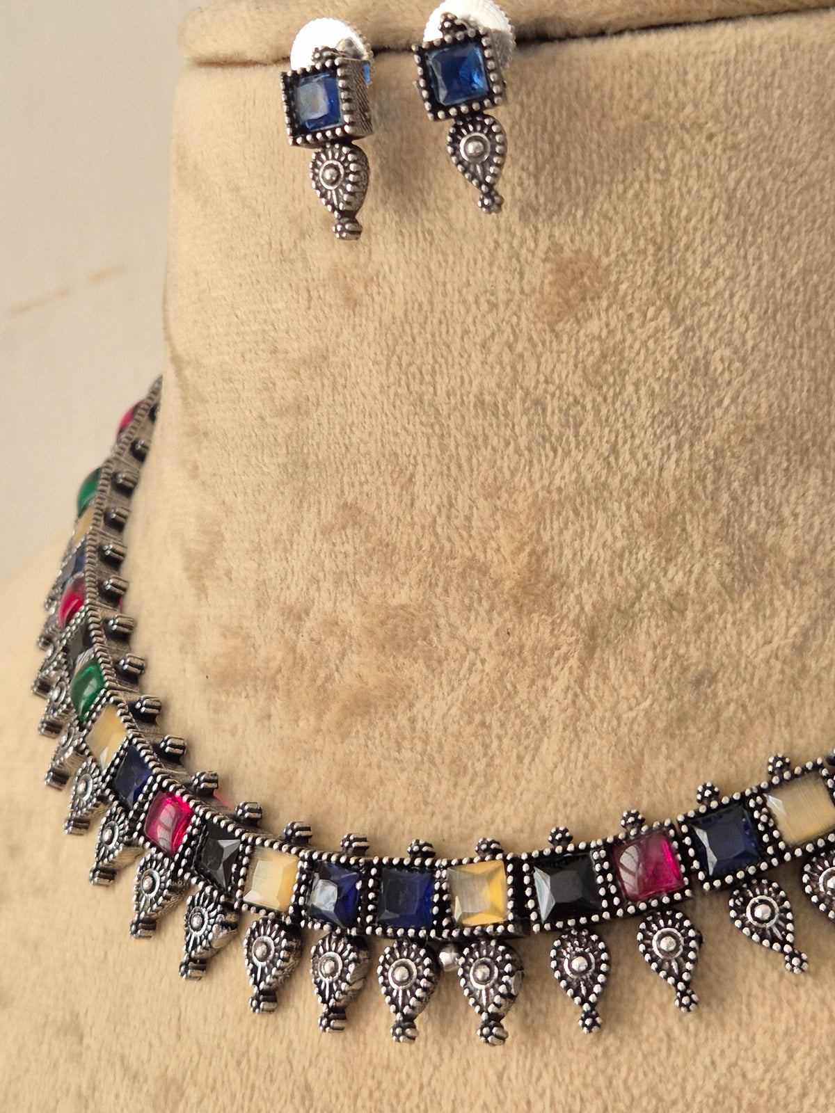 A silver-toned jewelry set featuring a choker necklace and matching drop earrings with rectangular multicolored gemstones and dangling ornate silver motifs by naitri.in