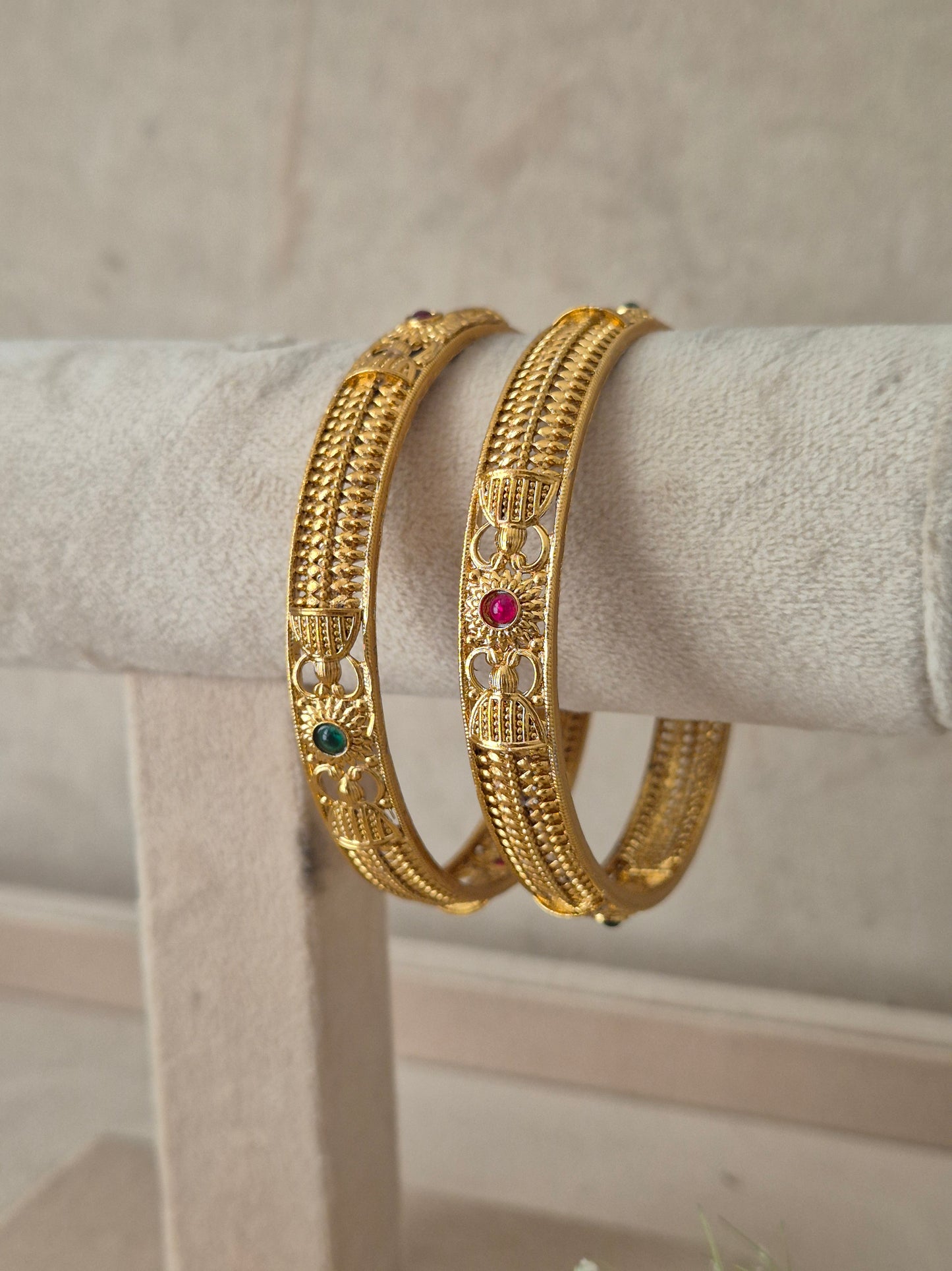 A pair of gold-plated Indian bangles with intricate filigree work and small red and green stone accents, by naitri.in