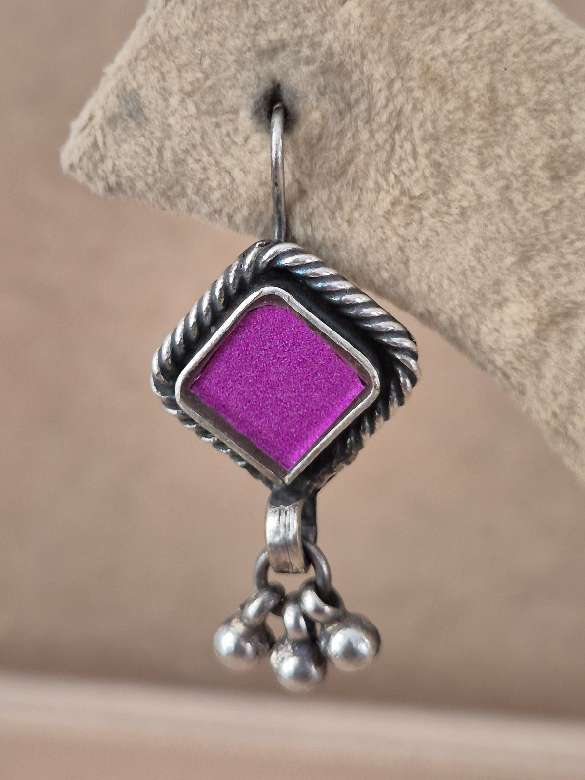 A close-up of a silver dangle earring featuring a textured, twisted border and a bright purple square center, hanging from a silver fishhook by naitri.in