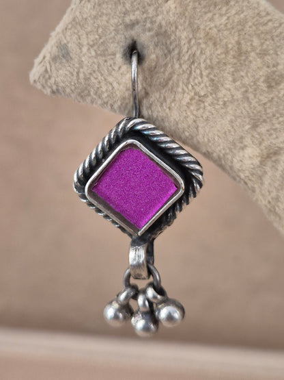 A close-up of a silver dangle earring featuring a textured, twisted border and a bright purple square center, hanging from a silver fishhook by naitri.in
