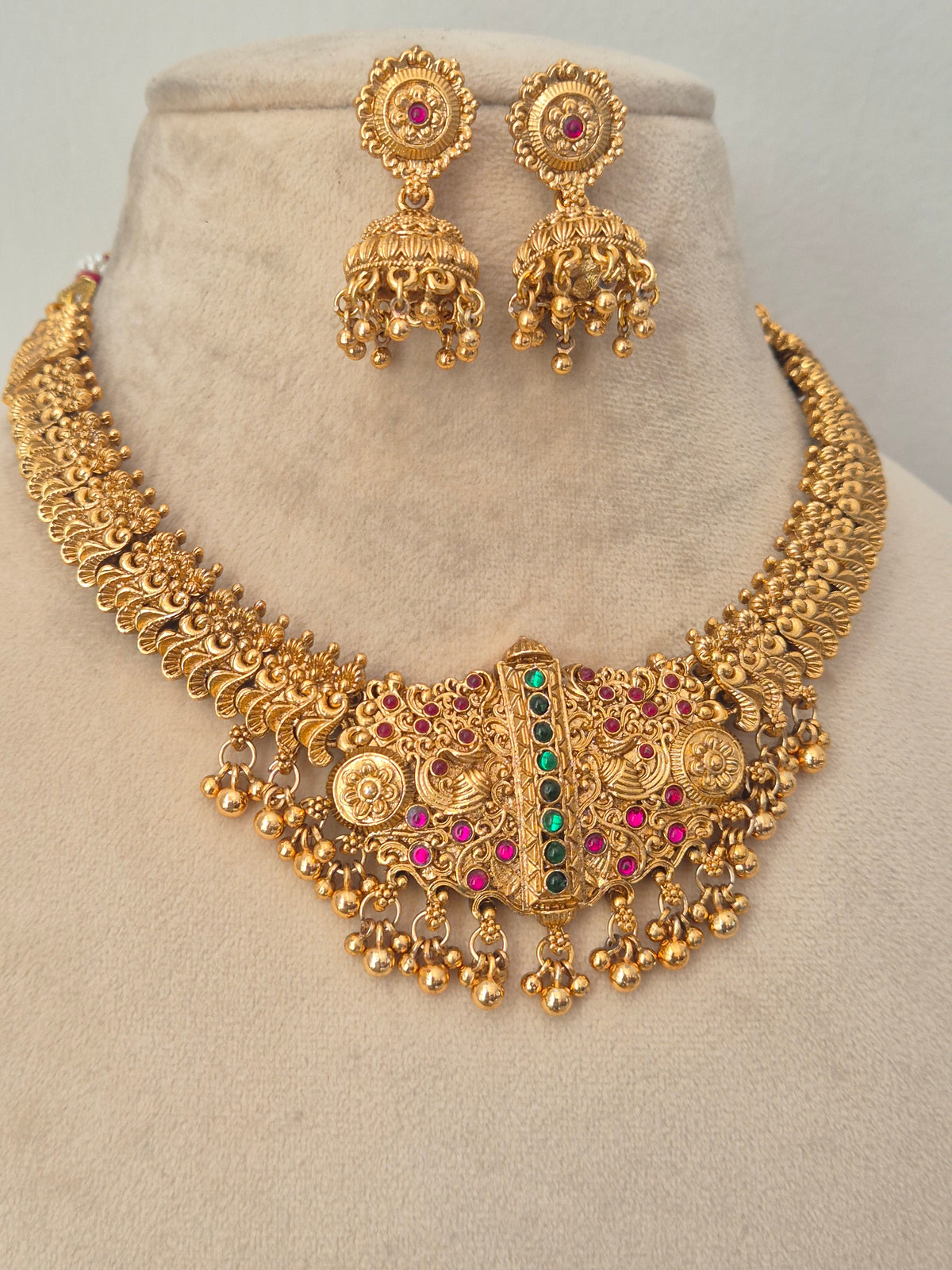 An ornate gold-toned statement necklace features intricate filigree detailing, multiple dangling coin-like elements, and a central vertical bar set with green stones and small pink stones by naitri.in