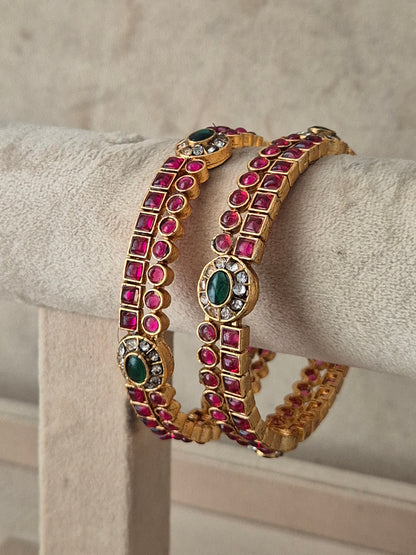 A set of two gold-plated bangles decorated with rows of pinkish-red square gemstones and oval green stone accents, by naitri.in