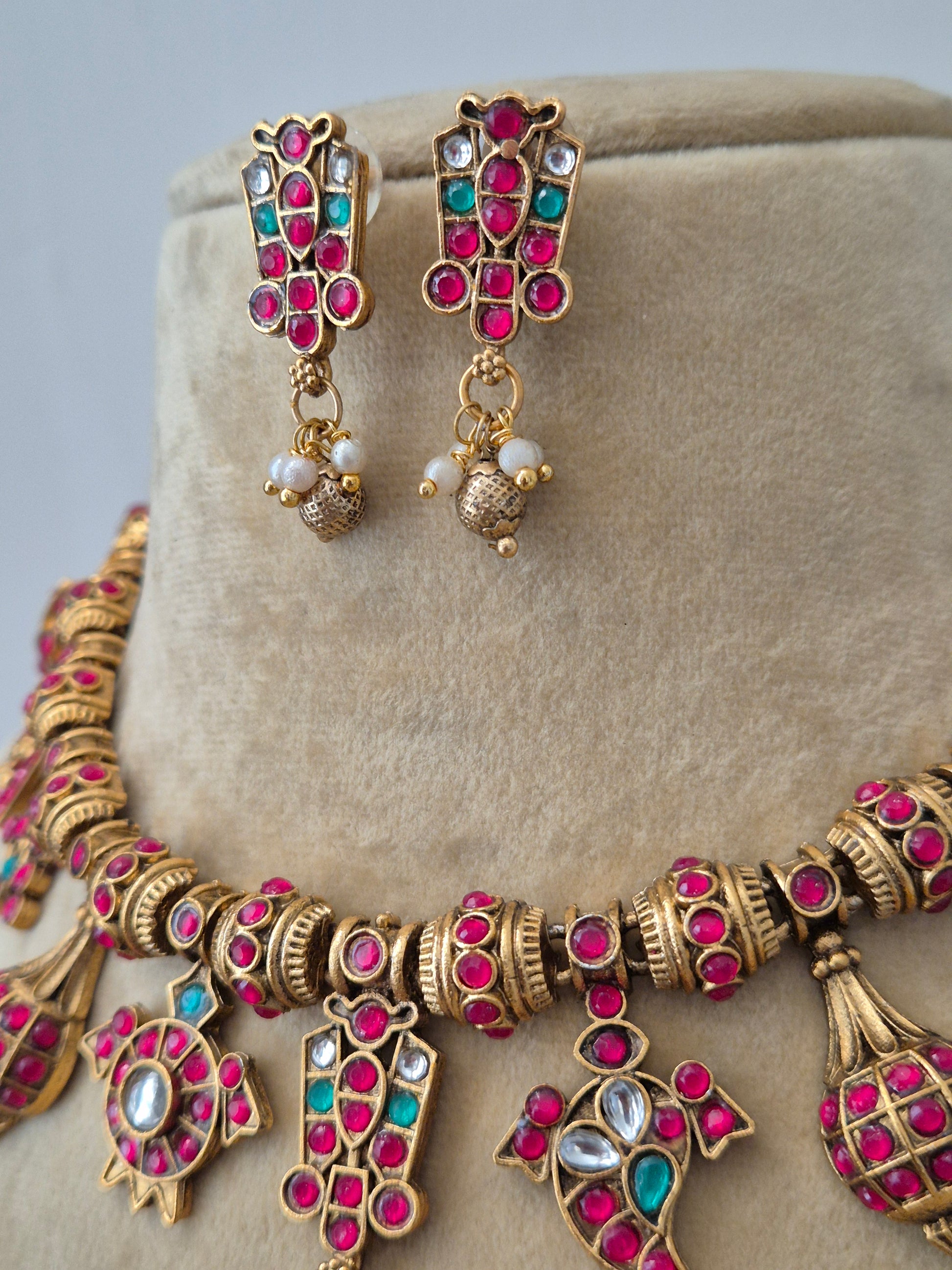 Traditional Indian imitation jewelry set showing a brass alloy gold necklace studded with pink and green gemstones, tiny pearls, and matching ethnic earrings by naitri.in