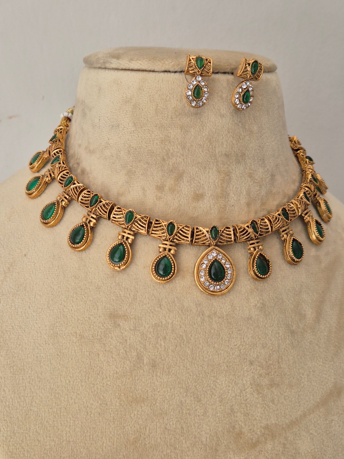Gold-toned traditional jewelry set including a necklace and matching earrings, decorated with green teardrop-shaped stones and small clear stones by naitri.in