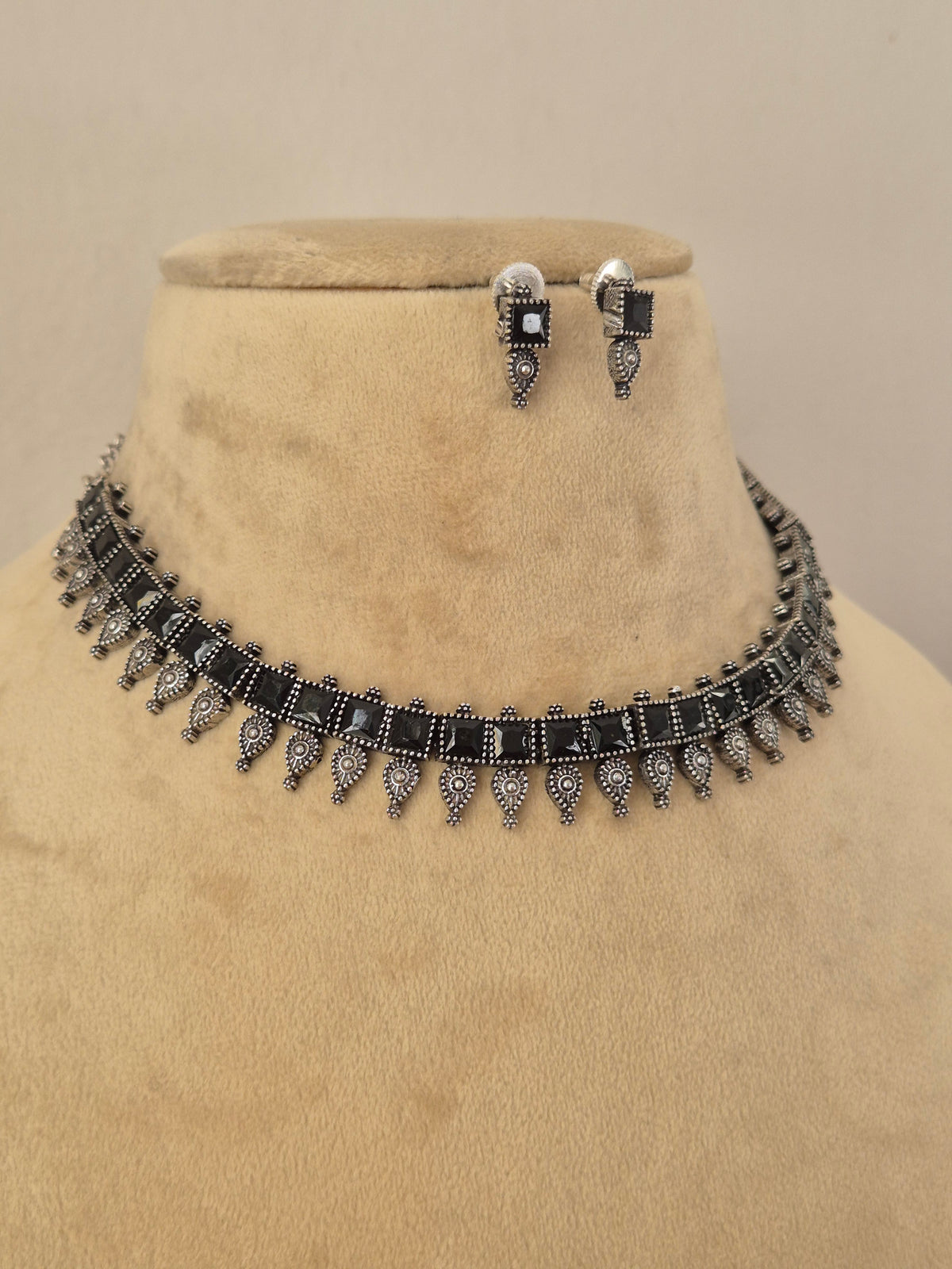 A silver-toned jewelry set featuring a choker necklace and matching drop earrings with rectangular black gemstones and dangling ornate silver motifs by naitri.in