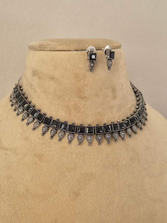 A silver-toned jewelry set featuring a choker necklace and matching drop earrings with rectangular black gemstones and dangling ornate silver motifs by naitri.in