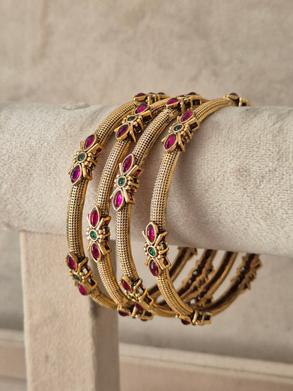 A stack of four gold-toned bangles with textured bands and small floral motifs set with pink and green stones, by naitri.in