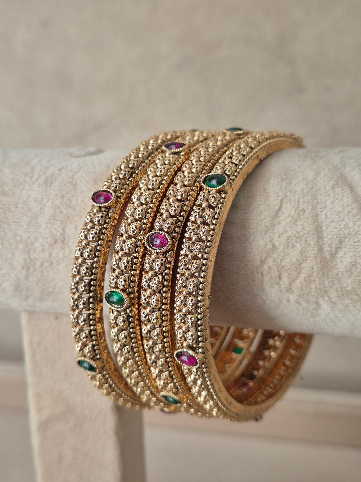 A stack of four gold-toned bangles adorned with clear stones and small green and pink gemstone accents, by naitri.in
