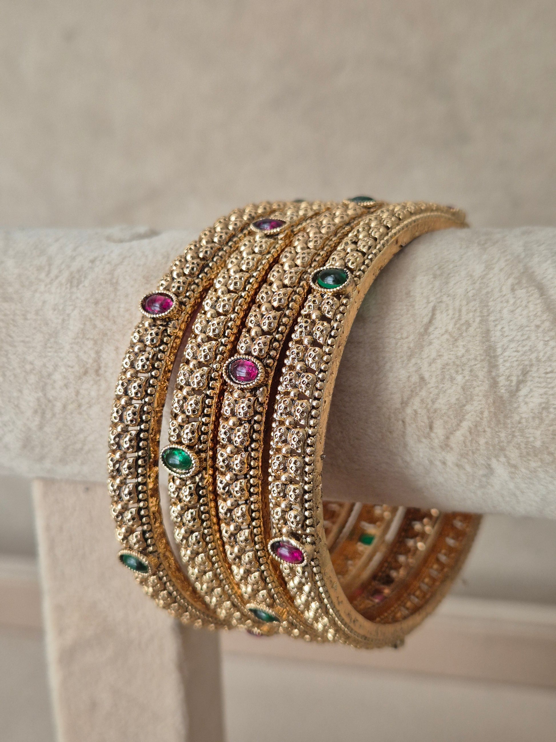 A stack of four gold-toned bangles adorned with clear stones and small green and pink gemstone accents, by naitri.in