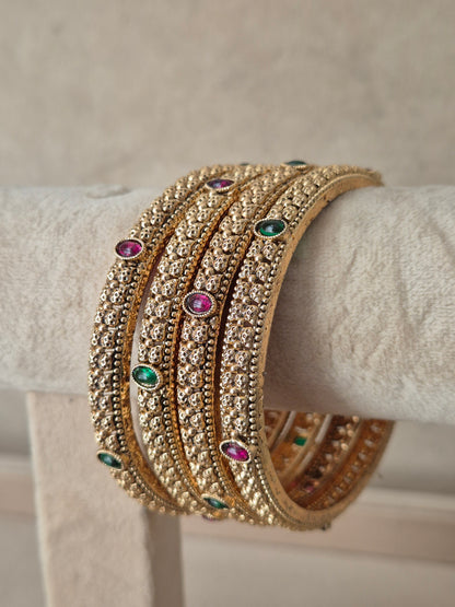 A stack of four gold-toned bangles adorned with clear stones and small green and pink gemstone accents, by naitri.in