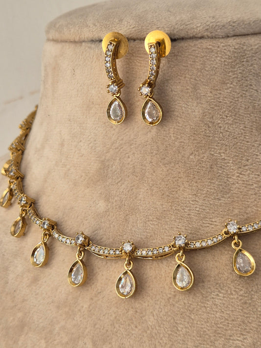 Gold jewelry set featuring a necklace and matching drop earrings, both adorned with clear teardrop-shaped gemstones and small clear stones along the curved bands by naitri.in