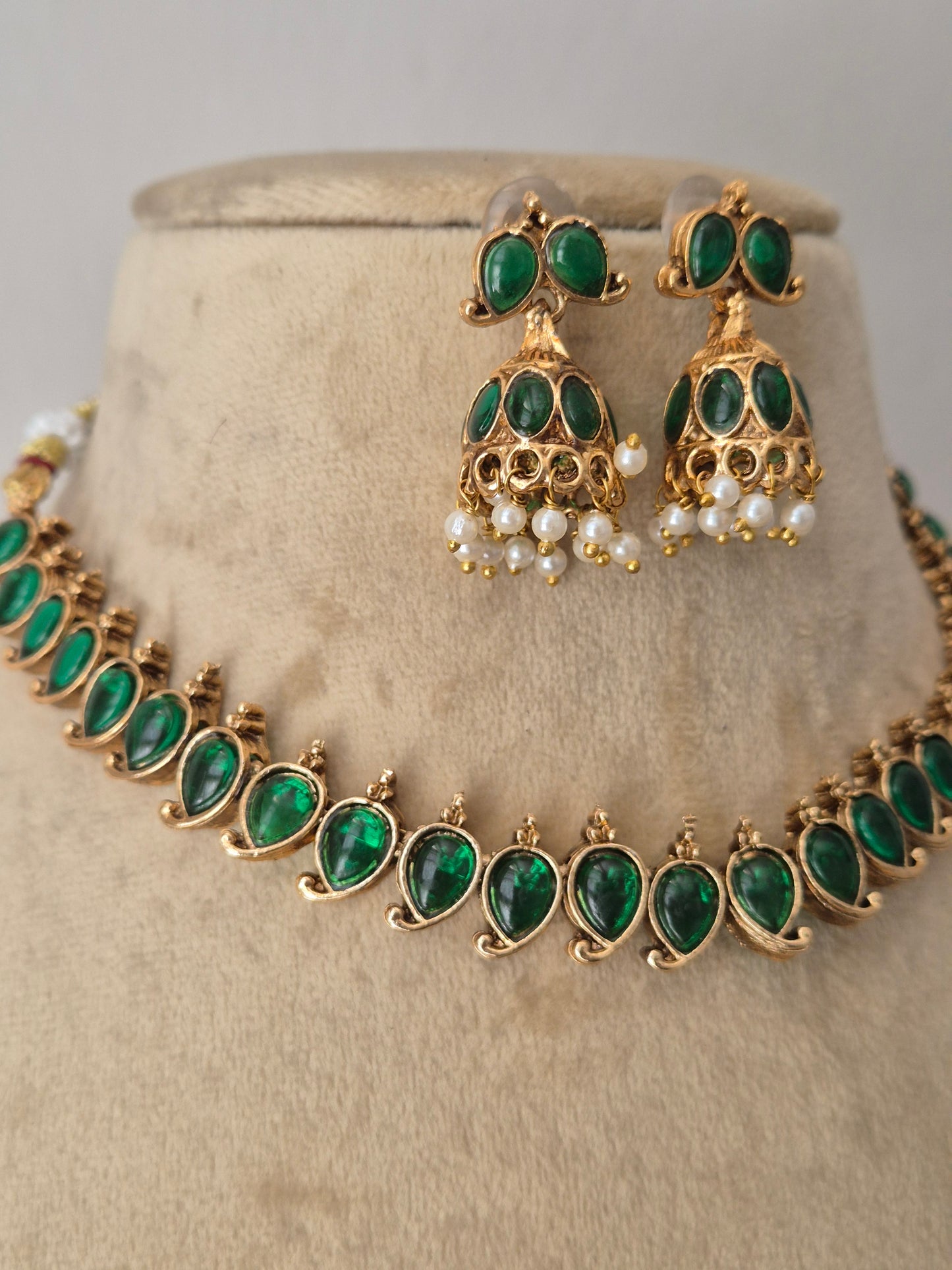 Gold-toned jewelry set with emerald green teardrop stones and small white pearl accents. The set includes a necklace and matching earrings. The choker features a row of vibrant oval-cut green gemstones bordered by tiny gold accents, paired with matching bell-shaped jhumka-style earrings embellished with green stones and hanging white pearl droplets by naitri.in