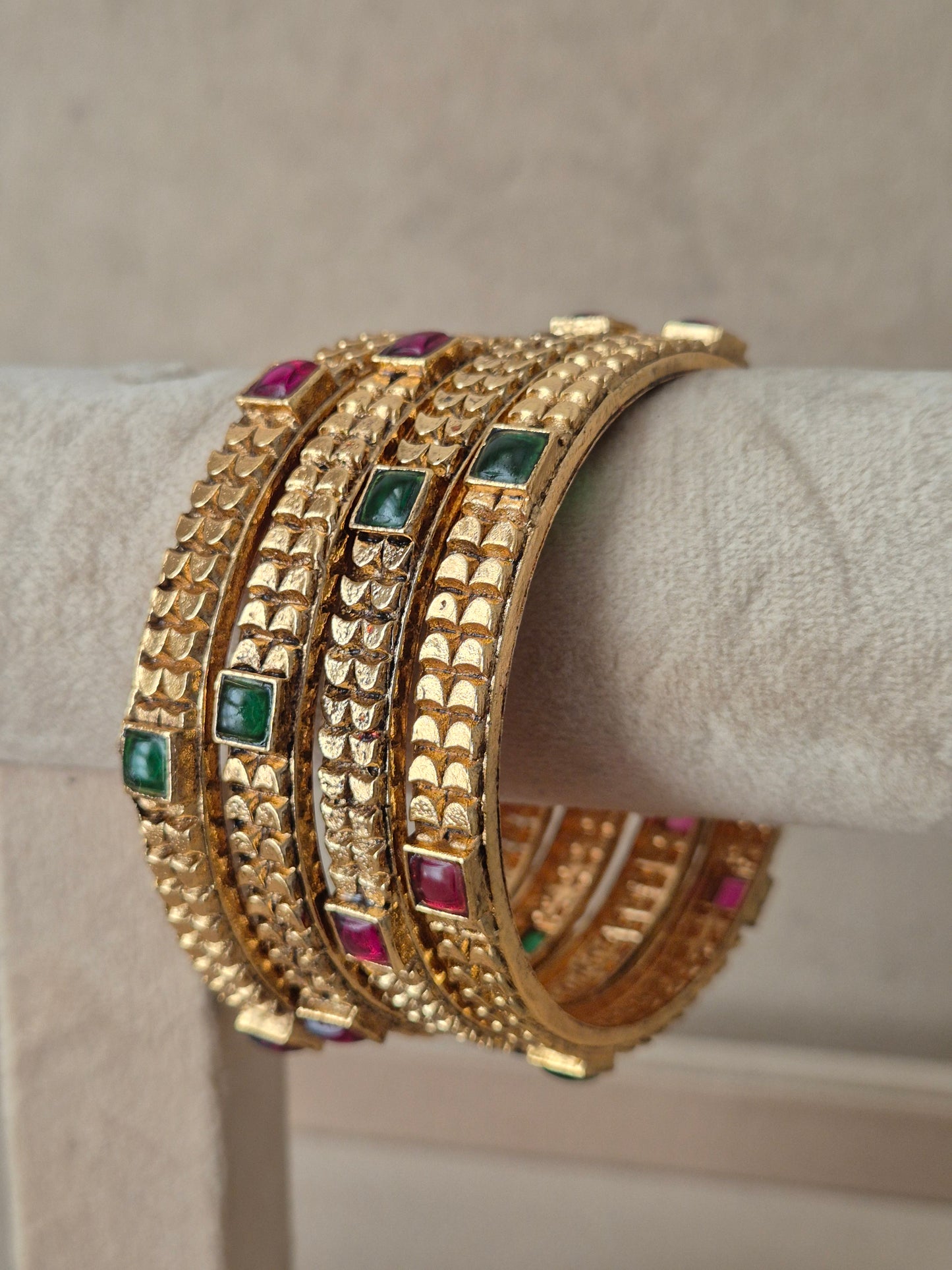 A stack of four gold-plated bangles with textured fish-scale patterns and small square green and pink gemstone accents by naitri.in