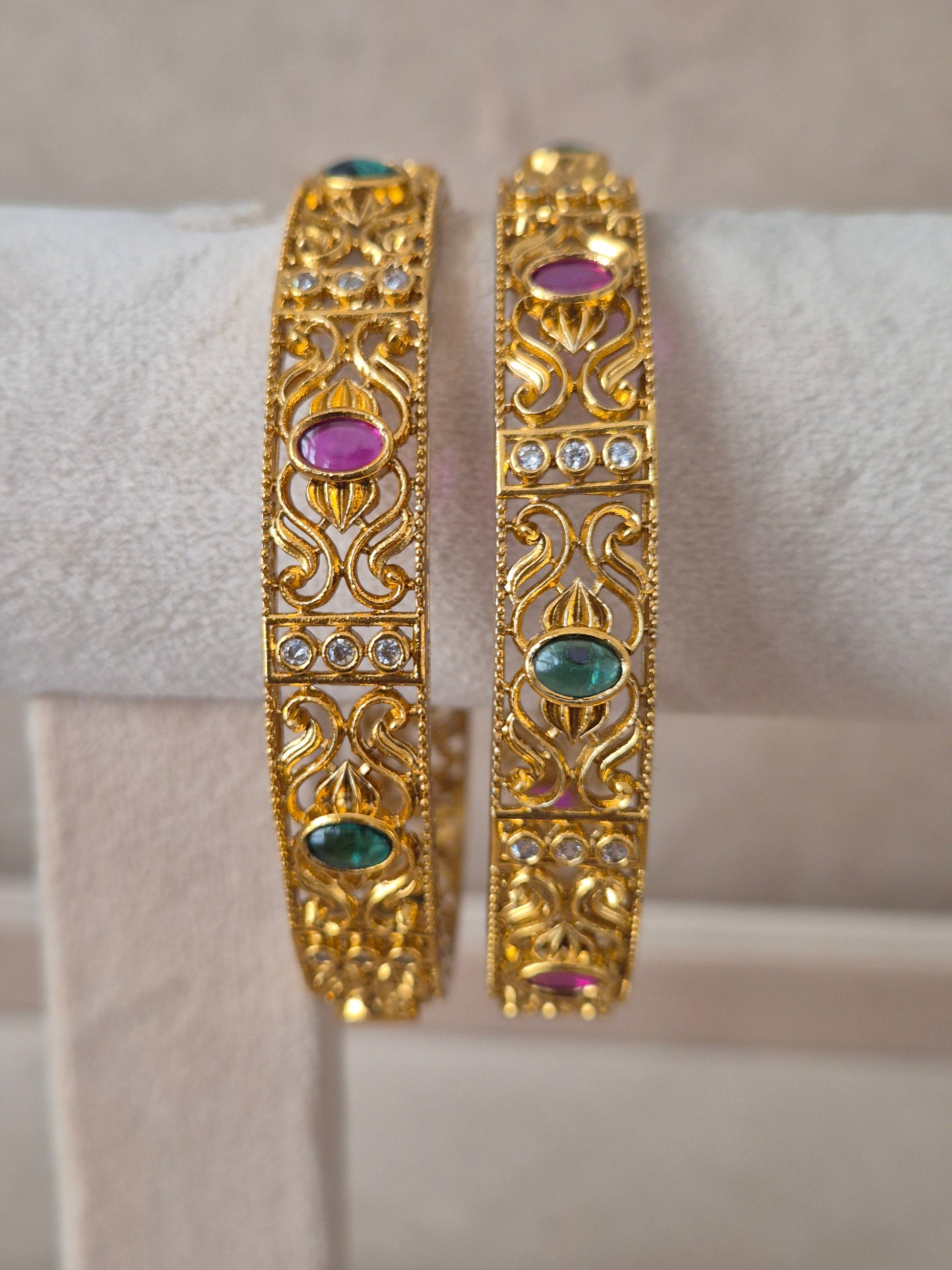 Pair of Naitri's Nagma gold bangles showcasing detailed ethnic craftsmanship, embedded with vibrant pinkish-red and green accent stones, by naitri.in
