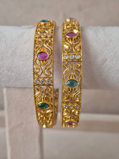 Pair of Naitri's Nagma gold bangles showcasing detailed ethnic craftsmanship, embedded with vibrant pinkish-red and green accent stones, by naitri.in