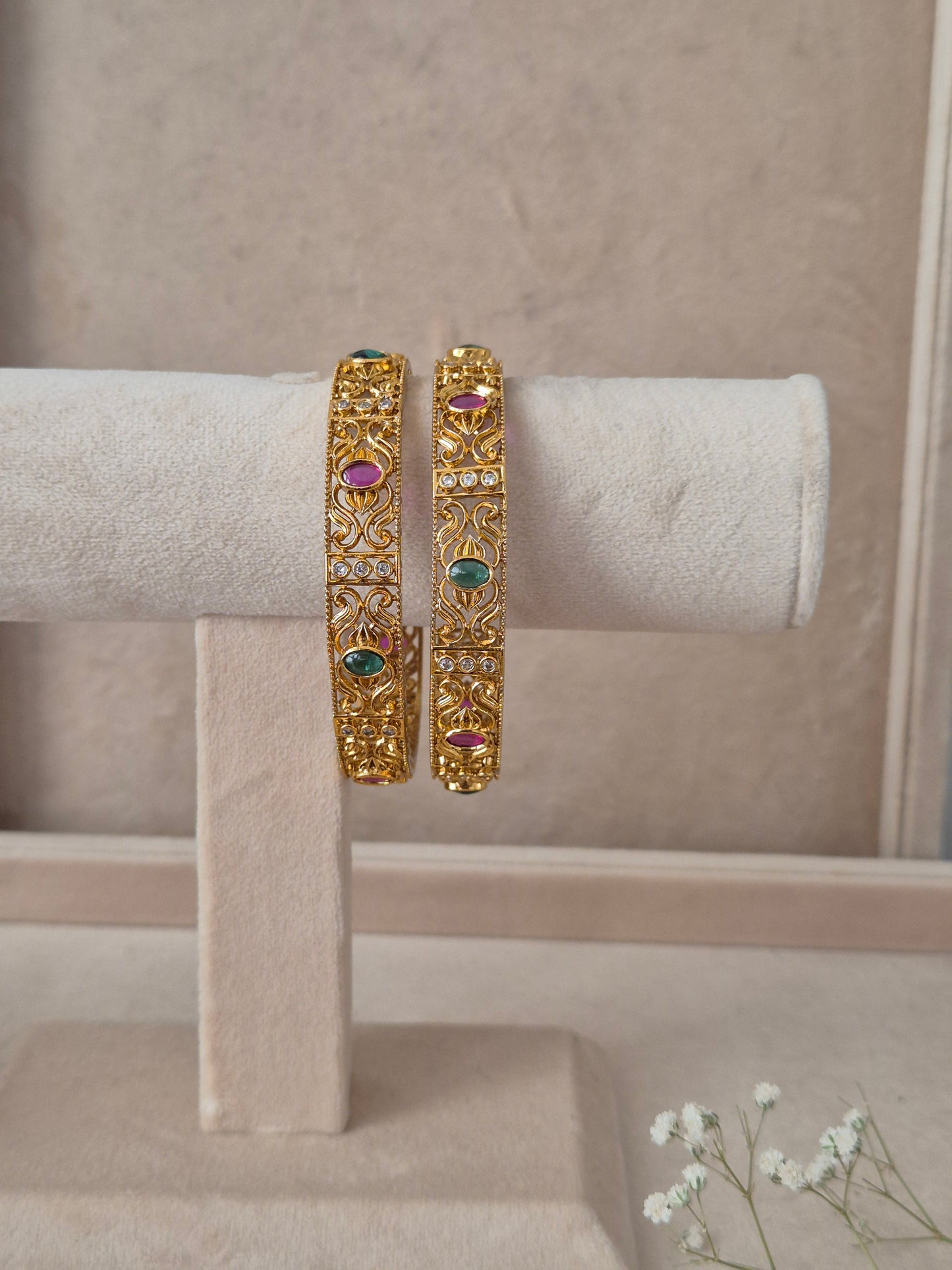 Set of 2 Nagma gold bangles by Naitri featuring intricate traditional patterned carvings, accented with alternating ruby-red and emerald-green gemstones by naitri.in