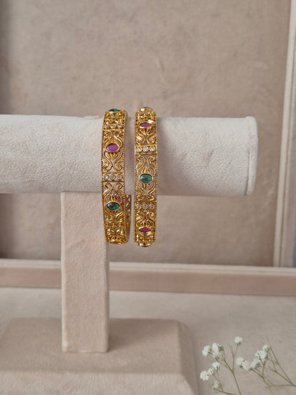 Set of 2 Nagma gold bangles by Naitri featuring intricate traditional patterned carvings, accented with alternating ruby-red and emerald-green gemstones by naitri.in