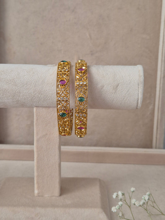Set of 2 Nagma gold bangles by Naitri featuring intricate traditional patterned carvings, accented with alternating ruby-red and emerald-green gemstones by naitri.in