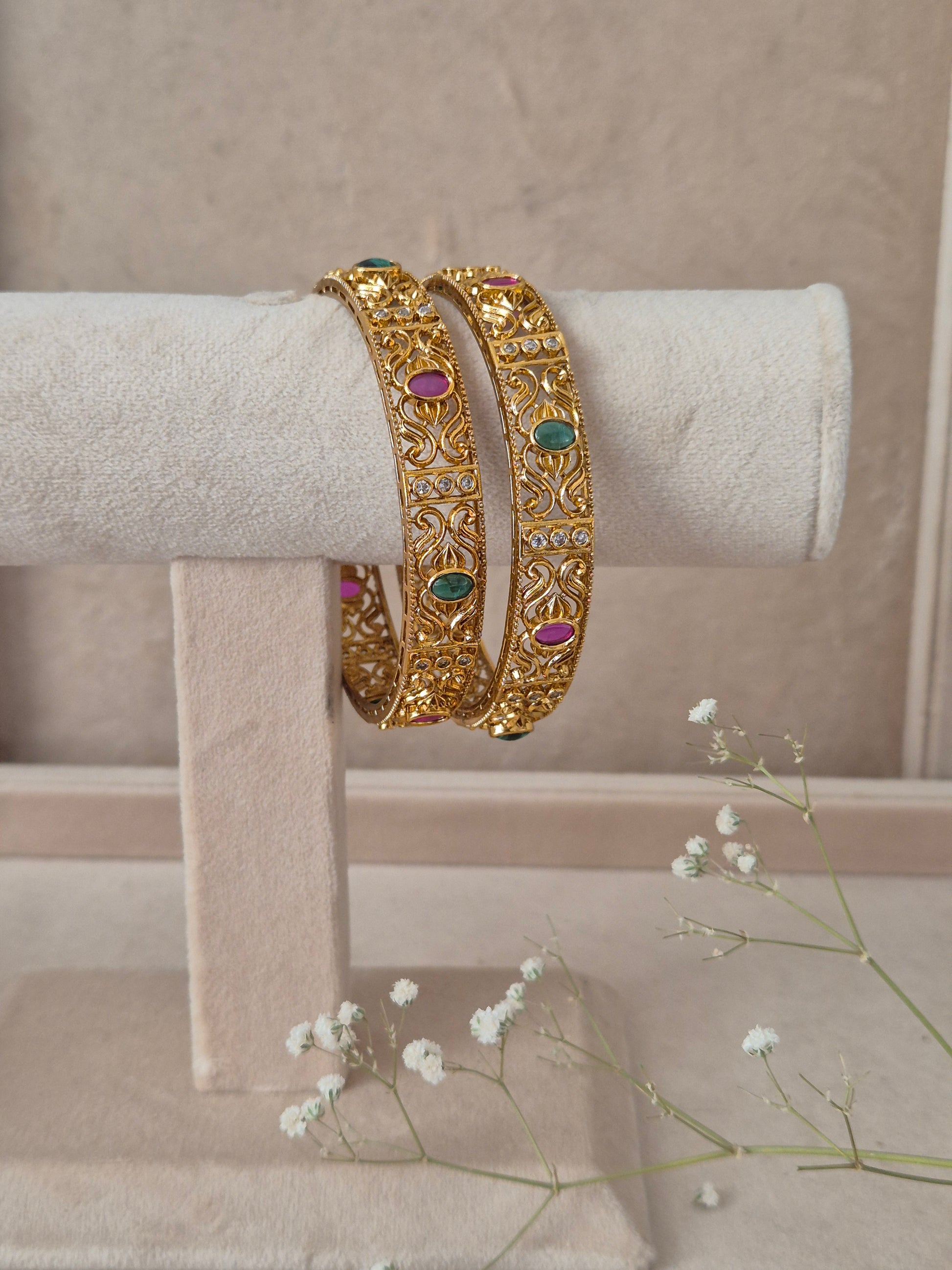 Two traditional gold-plated ethnic bangles with detailed geometric engravings and inlaid red and green gemstones