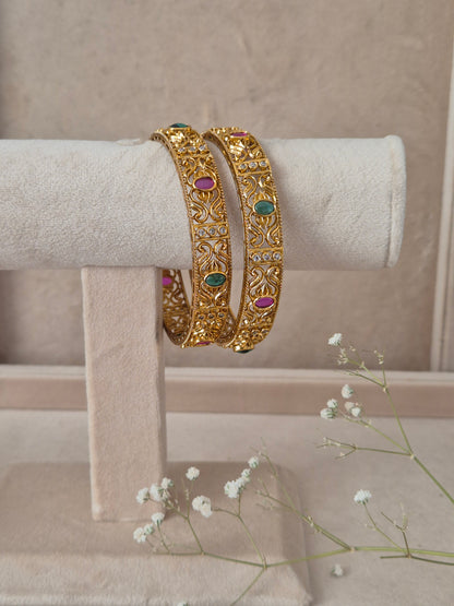 Two traditional gold-plated ethnic bangles with detailed geometric engravings and inlaid red and green gemstones
