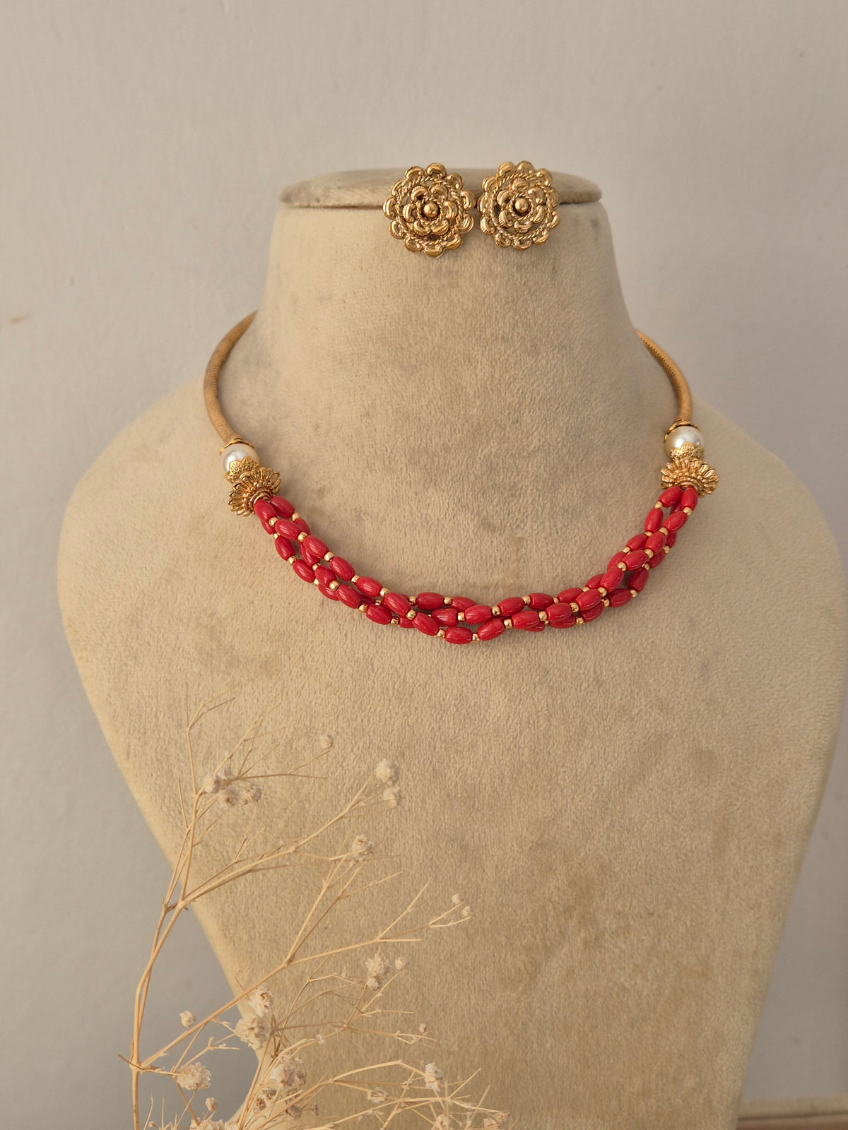 Handcrafted Golden Ethnic Necklace with bright red beaded strands styled on a display bust, accompanied by matching floral-textured gold-toned studs by Naitri.in