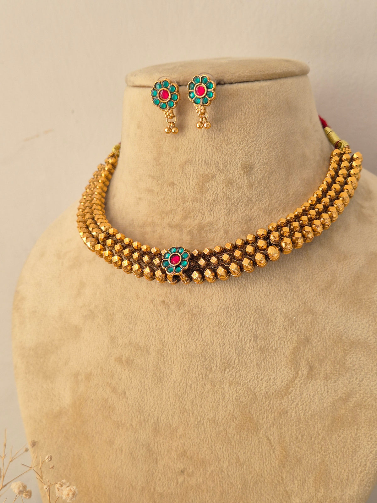 Traditional Indian gold-toned multi-strand bead necklace with a floral centerpiece featuring green and pink stones, alongside matching stud earrings by Naitri.in