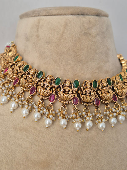 Traditional Indian gold-plated choker necklace and jhumka earrings set featuring intricate temple motifs with ruby and emerald stone embellishments, accented with delicate hanging pearl drops by Naitri.in