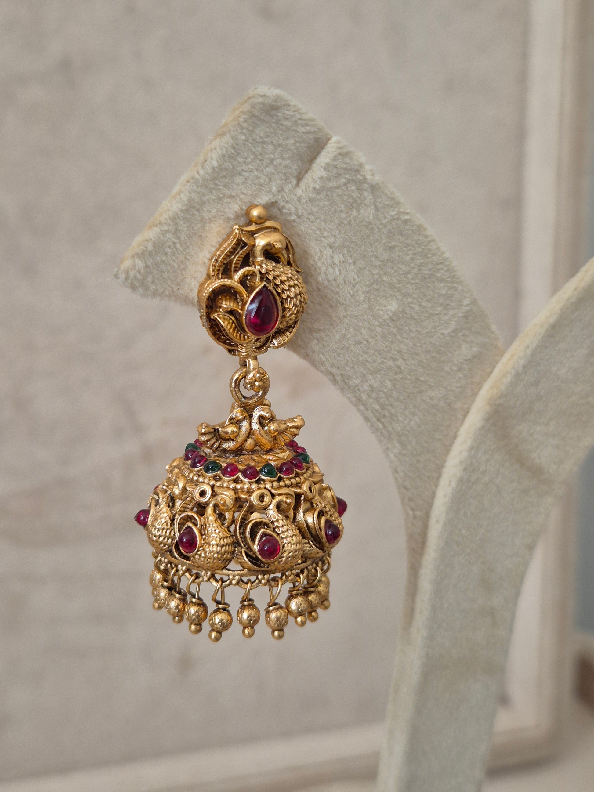 Handcrafted antique gold jhumka earrings with a teardrop pink stone stud, peacock engravings, and delicate metallic gold bead droplets by naitri.in