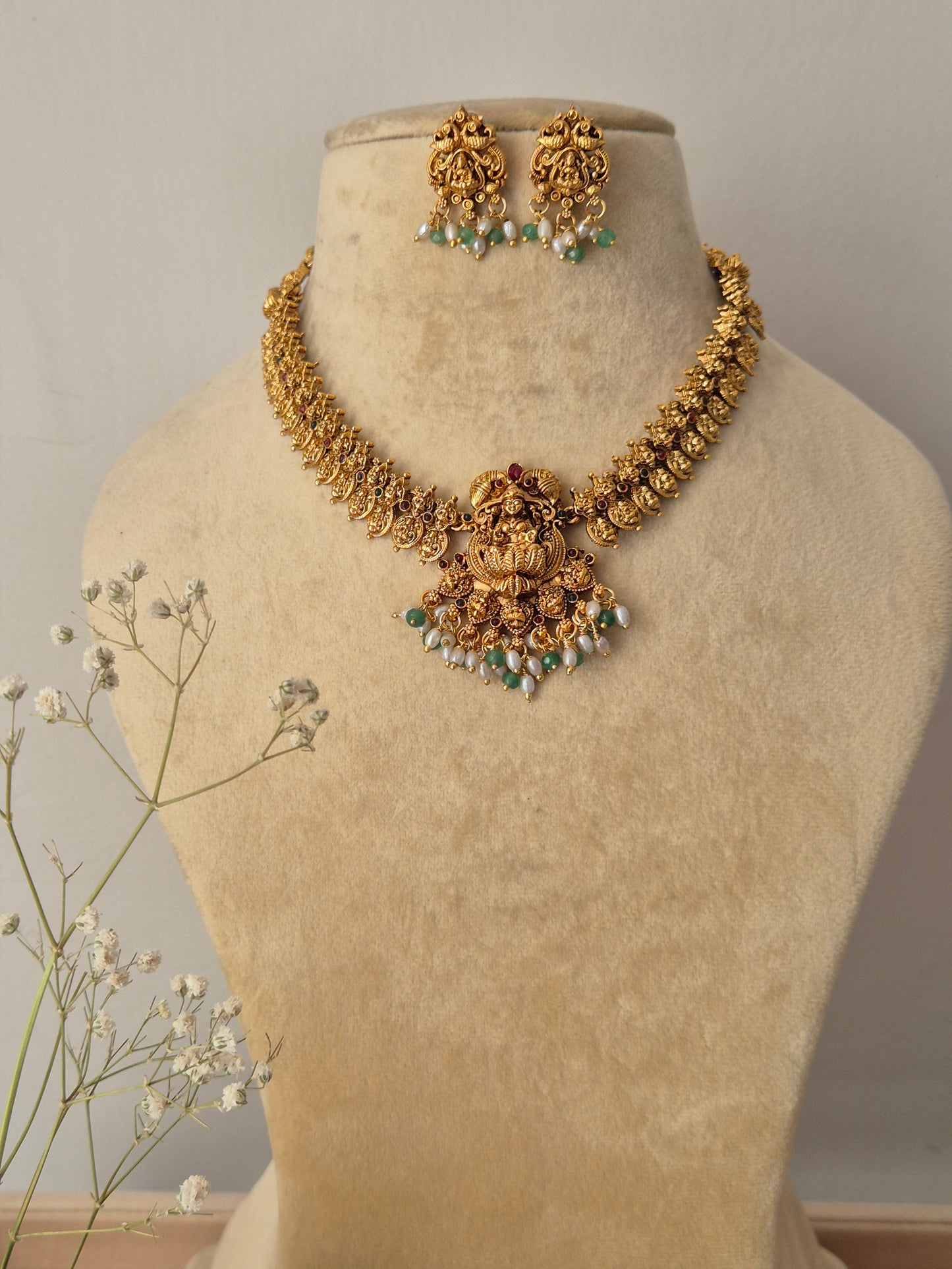 Handcrafted traditional gold-plated temple jewelry necklace featuring an intricate Goddess Lakshmi motif pendant with green beads and pearl cluster fringes, paired with matching stud earrings By naitri.in